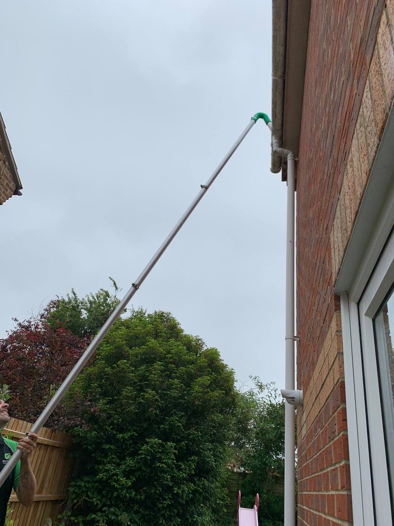 A person using a long pole gutter vacuum system to clean a high gutter on a brick house. The pole extends upward to reach the gutter, with trees and a cloudy sky in the background.