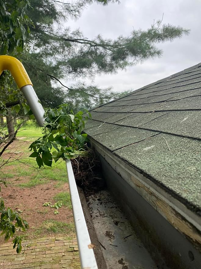 A close-up view of a house roof with green asphalt shingles and a white gutter filled with dirt and plant growth. A yellow-handled gutter cleaning tool is visible, pulling out leafy debris.